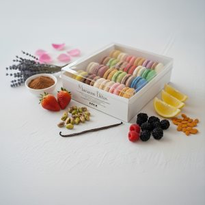 A box of Macarons Strawberry and Vanilla, 24 count (Copy), is surrounded by strawberries, blackberries, raspberries, lemon slices, almonds, pistachios, lavender, cocoa powder, and vanilla bean on a white surface.