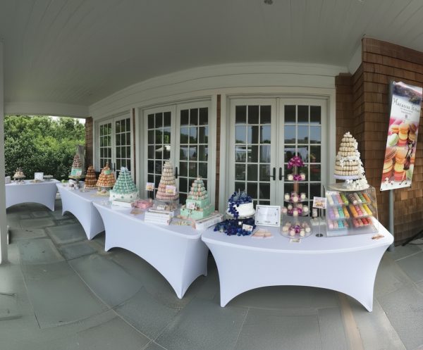 A row of tables covered with white cloths displays Macarons Strawberry and Vanilla, 24 count, and pastry towers in front of glass doors on a shaded patio, with a macaron poster on the right.